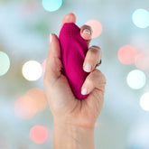 A hand holds the OhMiBod Foxy Bluetooth Panty Vibrator, a small, pink, textured object against a blurred background with colorful bokeh lights. Its oval shape features Velvet Wave technology on one end, providing a unique tactile experience.